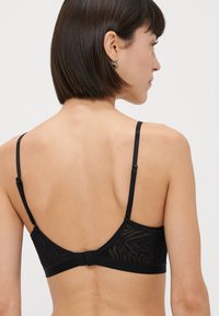 Ženska s kratkimi temnimi lasmi nosi črn čipkast modrček z nastavljivimi naramnicami, prikazana zadaj proti enostavni ozadju.