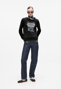 Svart genser med grafisk design foran, blå denim straight-leg bukser og svart-hvite joggesko. Tilbehør inkluderer solbriller og et skjerf.