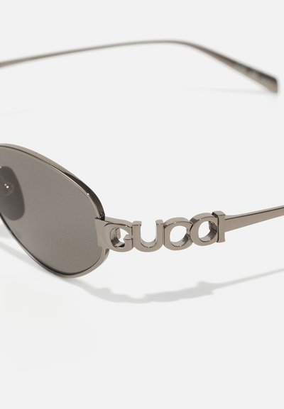 Ochelari de soare cu lentile întunecate, ramă metalică subțire și un logo argintiu proeminent pe sprijin. Formă ovală cu o finisare netedă.