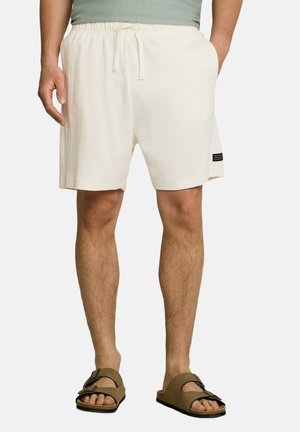 Hombre con pantalones cortos texturizados color marfil con cintura de cordón y sandalias marrones con hebilla, de pie frente a un fondo liso.