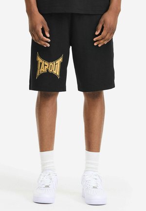 Czarne bawełniane szorty z dużym złotym logo "TAPOUT" na lewej nogawce, o luźnym kroju. Połączone z białymi trampkami i skarpetkami.