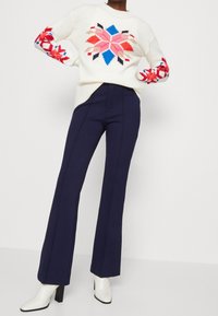 Pull en tricot blanc avec des motifs floraux colorés sur les manches, associé à un pantalon bleu marine à coupe évasée et des bottines blanches à talons.