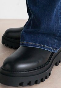 Svarte chunky-soled støvler med glatt finish, kombinert med mørke denimjeans som har en lett oppbrett på kanten. Grovt sålemønster.