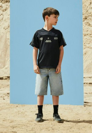 Pojke i svart sporttröja och jeansshorts står på sand och tittar åt sidan mot en ljusblå bakgrund och sandigt landskap.