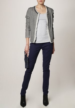 Femme portant une veste grise sur une chemise gris clair, un pantalon bleu marine, des bottines noires, tenant une pochette foncée.