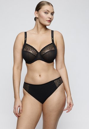 Woman wearing a black lingerie set with sheer patterned cups and matching high-waist panties, standing against a plain light background.