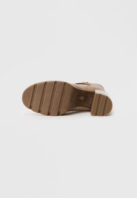 Botte de cheville beige avec un talon empilé, semelle en caoutchouc avec des rainures et des accents métalliques sur le côté. Tige texturée avec des détails de couture.