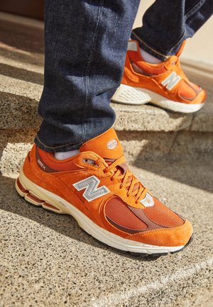 Osoba nosząca jaskrawo pomarańczowe sneakersy New Balance 2002R i ciemnoniebieskie dżinsy, stąpająca po teksturowanych betonowych schodach w świetle słonecznym.