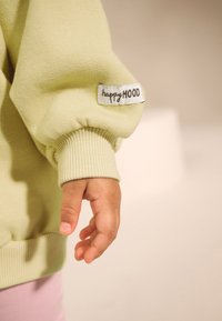 Light green sweater with ribbed cuffs, featuring a sewn label reading "happyMOOD" on the sleeve. Fabric appears soft and textured.