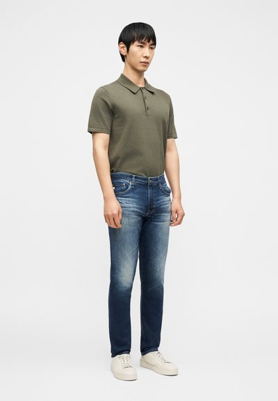 Άνδρας που στέκεται φορώντας ξεθωριασμένο μπλε τζιν, χακί πράσινη κοντομάνικη πόλο μπλούζα και λευκά αθλητικά παπούτσια, μπροστά σε απλό λευκό φόντο.