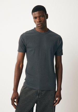 Giovane uomo con capelli corti che indossa una t-shirt aderente a maniche corte grigio scuro e jeans grigi scuri su uno sfondo chiaro e uniforme.