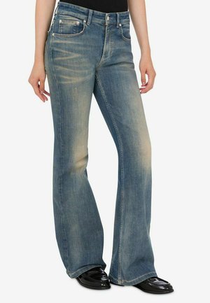Donna che indossa jeans svasati blu scoloriti e scarpe nere lucide, in piedi con le mani rilassate lungo i fianchi su sfondo bianco.