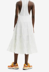 Vestido blanco de corte en capas con un diseño sin mangas y cierre trasero de botones. Combinado con zapatillas multicolores que tienen acentos en naranja y amarillo.
