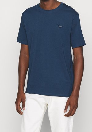 Mann trägt ein marineblaues T-Shirt mit kurzen Ärmeln und kleinem weißem Logo auf der Brust sowie weiße Hose, steht vor einfachem Hintergrund.