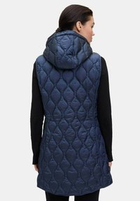 Námořnická modrá prošívaná vesta s kapucí, s texturovaným vzorem a mírně prodlouženým střihem. Zapínání na boční knoflík.