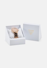Roségoldene Uhr mit rundem Ziffernblatt, Kristallakzenten und verlinktem Metallarmband, präsentiert in einer weißen strukturierten Box mit goldenem Logo.
