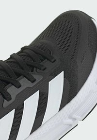 Musta urheilujalkine, jossa on teksturoitu kangasosa ja valkoisia yksityiskohtia, sekä kolme erottuvaa raitaa, suunniteltu tukemaan ja tarjoamaan mukavuutta.