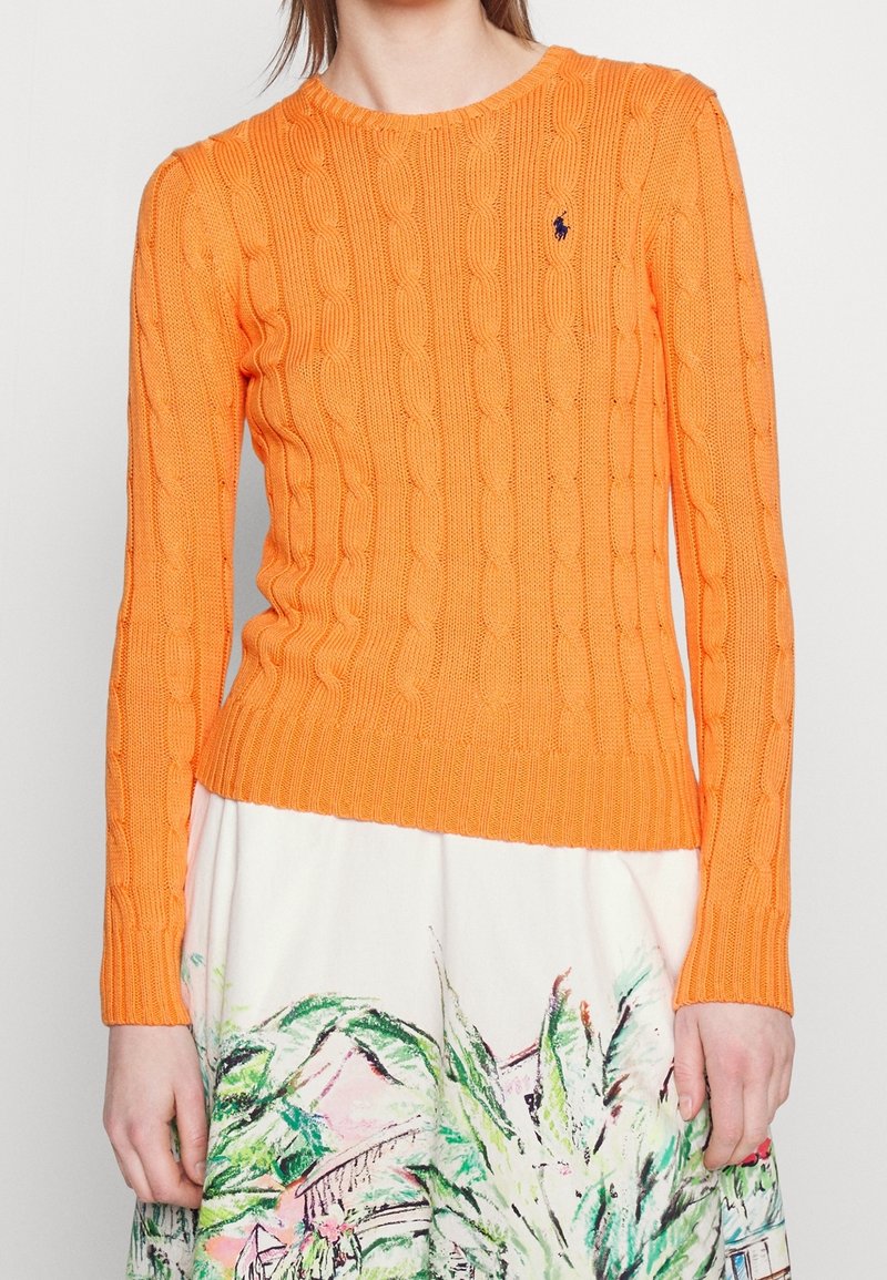Femme portant un pull en maille torsadée orange vif à manches longues et une jupe blanche avec des motifs botaniques abstraits verts et roses.