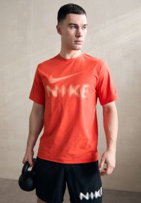 Punane spordipoliis, millel on suur beež Nike logo diagonaalis. Särgil on lühikesed varrukad ja ümmargune kaelaauk, koos mustade lühikestega, millel on valge tekst.