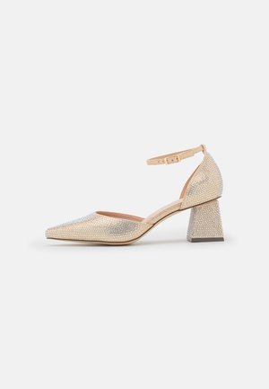 Chaussure dorée à bout pointu avec bride de cheville et talon bloc, recouverte de strass scintillants, vue de côté extérieur sur fond blanc.