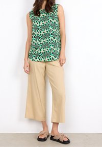 Ærmeløs grøn bluse med blomsterdesign, parret med beige løse bukser og brune sandaler med en knyttet detalje.