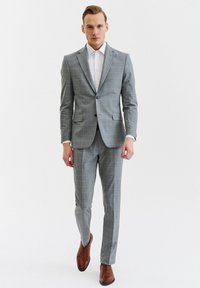 Szary garnitur w kratę z dwukluczową marynarką, klapami w kształcie notcha oraz dopasowanymi spodniami slim-fit. Brązowe skórzane buty dopełniają stylizację.