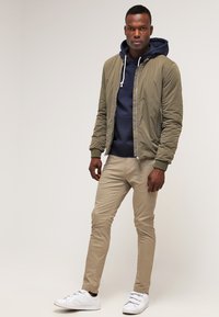 Olive green bomber jacket over a navy hoodie, paired with light beige trousers and white sneakers. Simple design, lightweight material.