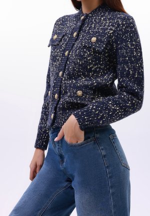 Marineblauwe trui met gouden spikkels, textuur breiwerk en twee borstzakken. Bevat een kraag en grote decoratieve knopen. Draagt met blauwe jeans.