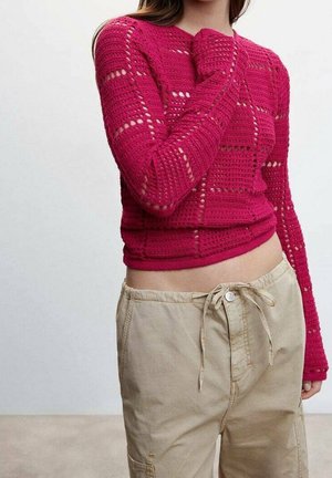 Femme portant un pull en maille rouge moulant à manches longues avec un pantalon beige à cordon, montrant le nombril et la main près du menton.