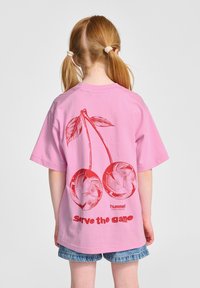 Rosa t-shirt med kort ärm som har en röd körsbärsgrafik och texten "serve the game" i fet röd bokstav. Tillverkad av bomull.
