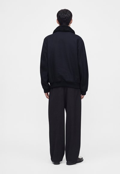 Homme debout, de dos, portant une veste noire avec un col en fourrure, un pantalon noir large et des chaussures noires sur un fond clair uni.