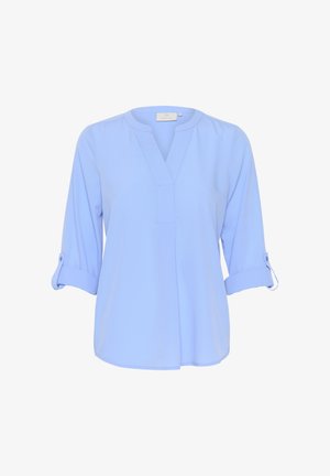 Gaiši zils, garo piedurkņu blūze no mīksta auduma. Ar V veida kakla izgriezumu, piedurkņu uzrullēšanu, ko nostiprina pogas, un brīvu piegriezuma formu.