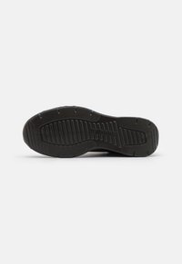 Schwarze Sport-Schuhsohle aus strukturiertem Gummi mit tiefen Rillen und Erhebungen für besseren Halt. Signatur-Logo in der Nähe der Ferse sichtbar.