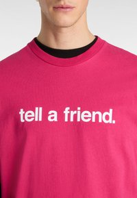 Lyserød bomulds t-shirt med rund halsudskæring og teksten "fortæl en ven." i store hvide bogstaver tværs over brystet.
