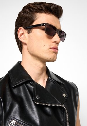Giovane uomo con gilet in pelle nero e occhiali da sole scuri che guarda di lato su sfondo bianco.