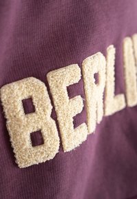 Burgundi szövet, textúrált krém „BERLIN” hímzéssel. A betűk kiemelkednek, puha, bolyhos felszínnel, amely kontrasztot képez a sima háttérrel.