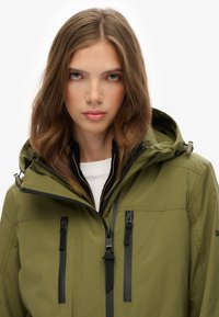 Chaqueta impermeable verde olivo con capucha, cremalleras negras y bolsillos laterales. Presenta una textura suave y un cordón ajustable alrededor de la capucha.