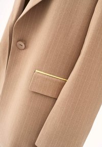 Béžové sako s tenkými pruhmi, jedným béžovým gombíkom a náprsné vrecko s úzkym vodorovným zlatým ozdobným prúžkom.