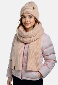 Ung kvinde iført en lys pink pufferjakke, en beige ribbet strikhue og et matchende langt tørklæde, smilende med den ene hånd i lommen.