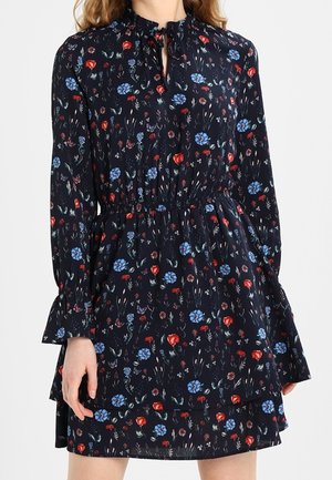Femme portant une robe noire à manches longues avec un imprimé floral bleu et rouge, taille cintrée, col à volants et jupe à étages.