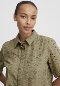 Short-sleeve, olive green shirt made of textured fabric, featuring floral cut-out patterns and a button-up collar.