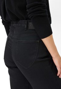 Jean noir taille haute avec une texture lisse, présentant une jambe fuselée, des poches arrière et une étiquette en cuir marquée au niveau de la taille.