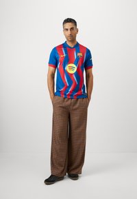 Mėlynos ir raudonos dryžuotos futbolo marškinėliai su apvaliu kaklu, derinami su plačiomis plaid kelnėmis rudos, smėlio ir tamsiai mėlynos spalvų, vilkint juodus batus.