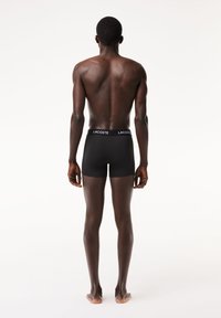 Fekete bokszernadrág, Lacoste logóval a derékrészen, testhez simuló fazon, sima textúra, hátulról látható egy meztelen bőrű modellen.