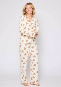 Two-piece pajama set in cream fabric featuring a teddy bear pattern. The top has long sleeves and a pocket; the pants are loose-fitting.