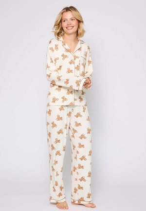 Two-piece pajama set in cream fabric featuring a teddy bear pattern. The top has long sleeves and a pocket; the pants are loose-fitting.