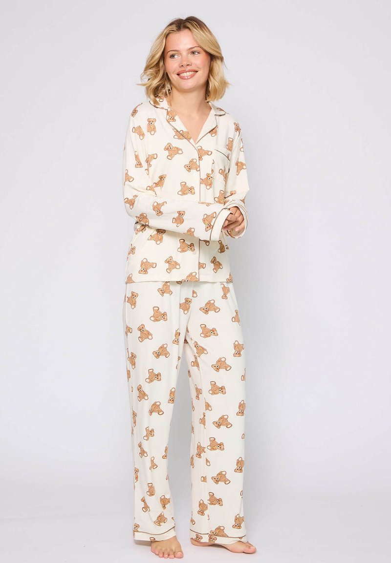 Two-piece pajama set in cream fabric featuring a teddy bear pattern. The top has long sleeves and a pocket; the pants are loose-fitting.