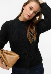 Czarny sweter na guziki o dzianinie w warkocze, z okrągłym dekoltem, sześcioma guzikami i taliowanym krojem. Połączony z trójkątną brązową skórzaną torebką.