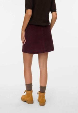 Vrouw in een donkerbruine top, bordeauxrode corduroy rok, grijze sokken en lichtbruine veterschoenen tot de enkel, staand tegen een effen witte achtergrond.