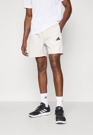 Homme portant un short blanc à motifs avec un logo noir, des chaussettes blanches avec un logo noir, des baskets noires et blanches, et un t-shirt blanc, debout.